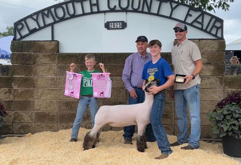 Cooper stands under the Plymouth County Fair sign, holding the head of his sheep. Also in the photo are another boy and two men.
