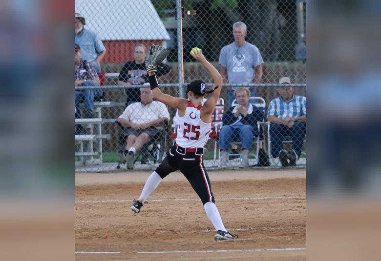 Gaswint throws a pitch from the pitcher's mound, her back to the camera. In the background, behind a chain link fence, spectators sit on lawn chairs.