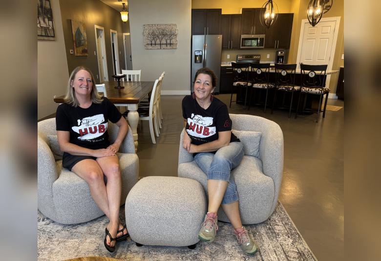 Stephanie Cacioppo and Jennifer Hart sit on stuffed chairs in the Airbnb living room. Both are wearing T-shirts that read "The HUBnb."