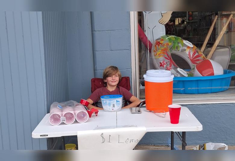 Oliver sits at a table outside of Dirks Hardware Hank. On the table are red solo cups and a large thermos.