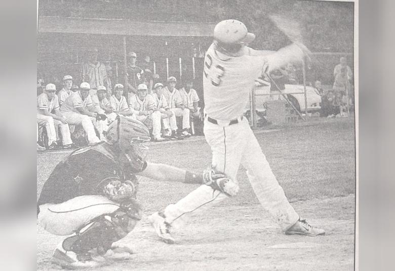 A black and white photo of a batter swinging, while the catcher stands behind them.