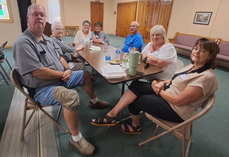 A half dozen people sit at a long wooden table, looking at the camera and smiling.