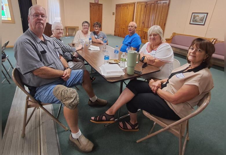 A half dozen people sit at a long wooden table, looking at the camera and smiling.