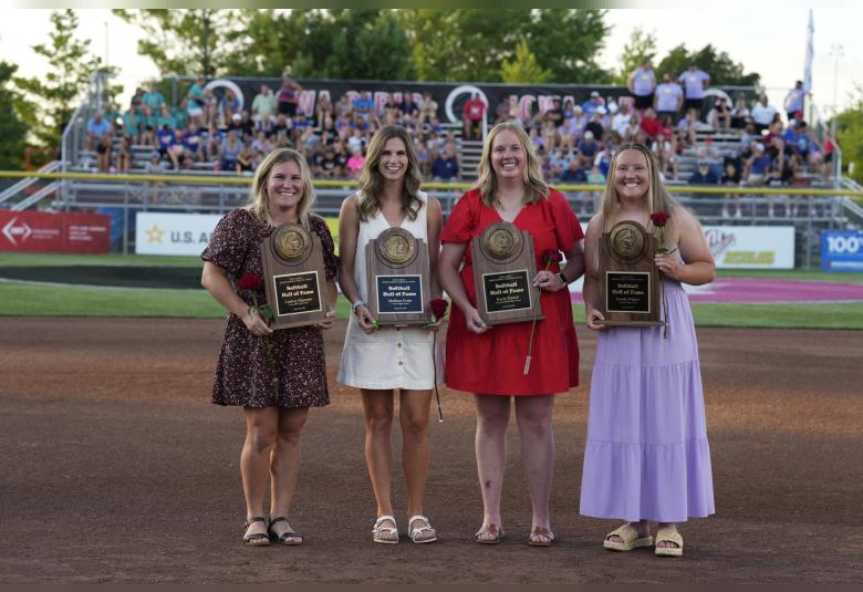 Four women stand outdoors, smiling and facing the camera. Each is holding a plaque that says "Softball Hall of Fame" and holding a red rose. In the background are spectators on bleachers.