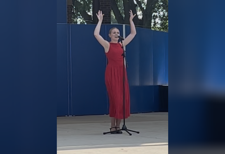 Kelsey stands on stage, wearing a red dress and singing into a microphone.