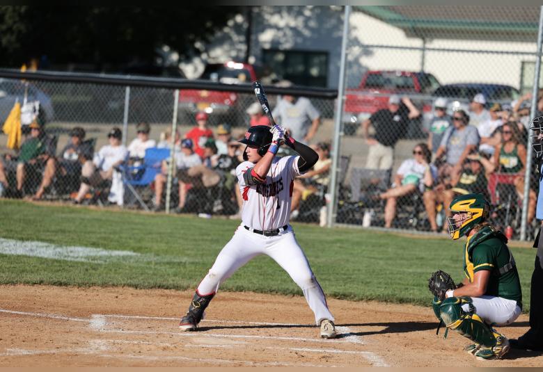 Hartman stands at bat, his body facing the camera and his turned to his right, facing the pitcher's mound. Behind him is the opposing team's catcher, who is crouched. In the background, spectators sit on lawn chairs behind a chain link fence.